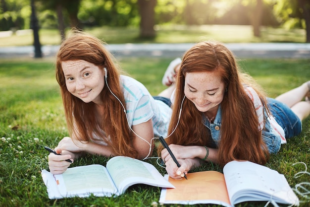 Homework are you kidding me Two charming girls with red hair lying and chilling on grass during free time doing homework sister helping sibling with lessons smiling and laughing Copy space
