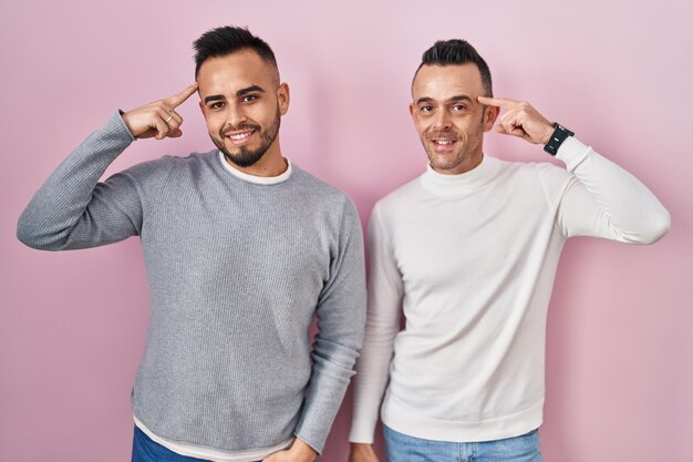 Гомосексуальная пара, стоящая на розовом фоне, улыбаясь, указывая пальцем на голову, отличная идея или хорошая память