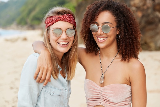 Homosexual family couple enjoy summer rest, have happy expression, embrace each other while stand against beach with mountain. People, friendship and same sex relationship concept