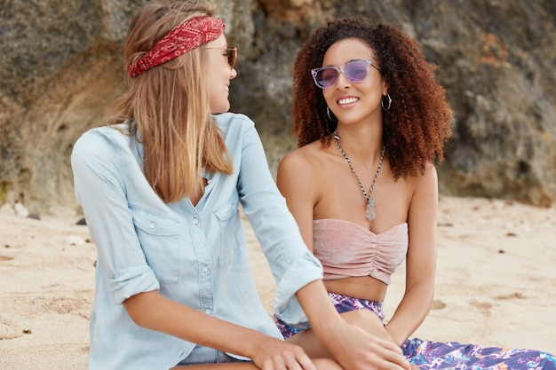 Homosexual mixed raced couple have romantic date outdoor at beach against cliff, have pleasant conversation, wear fashionable clothes and sunglasses, enjoy resort in hot country during summer