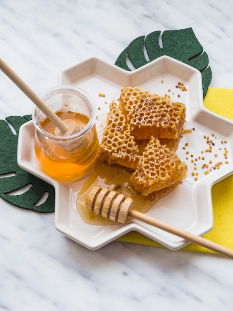 Honey dipper and honey comb with bee pollens in white tray on marble backdrop