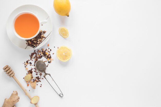 Honey dipper; strainer; herbs; lemon; ginger and cup of ginger tea on white backdrop