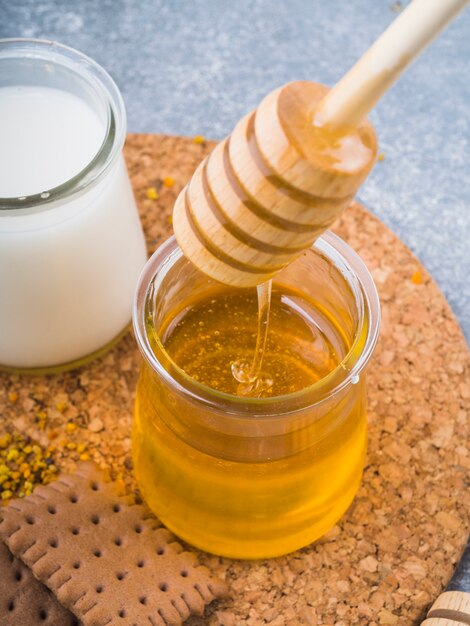 Honey dripping from dripper in glass pot with milk and biscuits on cork coaster