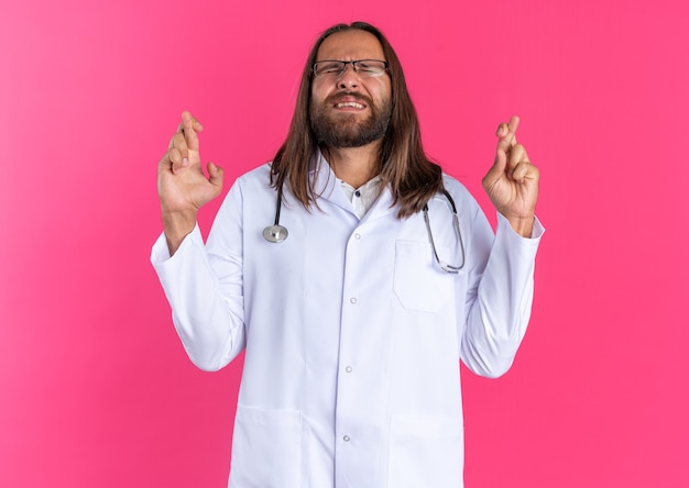 Hopeful adult male doctor wearing medical robe and stethoscope with glasses doing good luck gesture with closed eyes isolated on pink wall