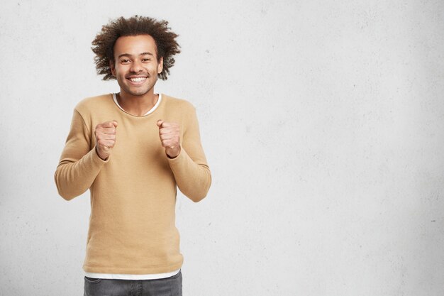 Hopeful Afro American male keeps hands in fists, smiles happily as waits for important decision