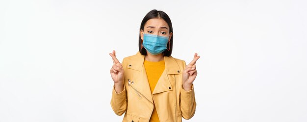 의료용 얼굴 마스크를 쓴 희망적인 아시아 소녀가 손가락을 교차시켜 smth 서 있는 것을 위해 기도하기를 바라고 있습니다.