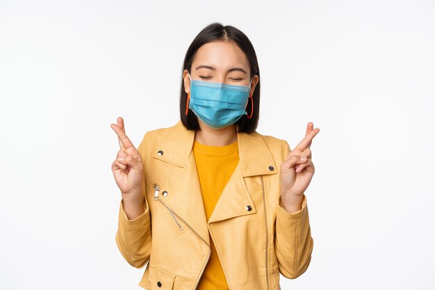 Hopeful asian girl in medical face mask cross fingers making wish hoping praying for smth standing with anticipation over white studio background