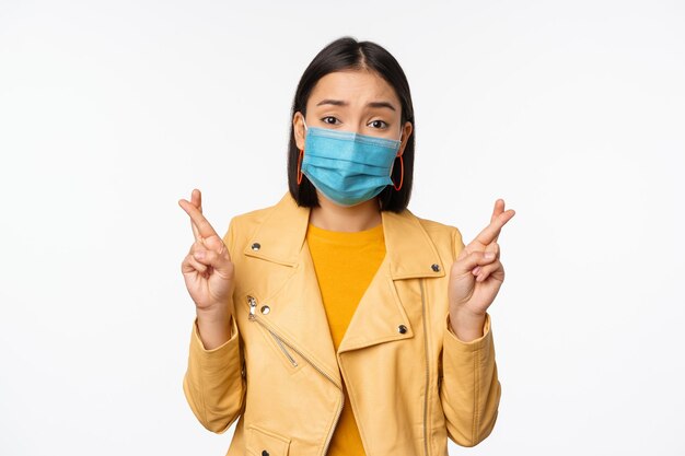 Hopeful asian girl in medical face mask cross fingers making wish hoping praying for smth standing with anticipation over white studio background