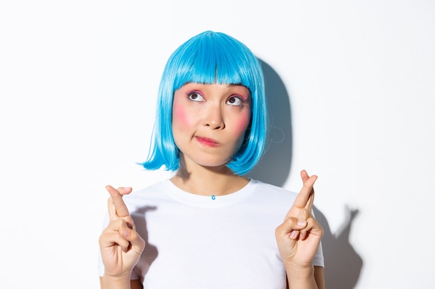 Foto gratuita speranza bella ragazza asiatica in parrucca blu che esprime desiderio, guardando sognante nell'angolo in alto a sinistra e incrociare le dita