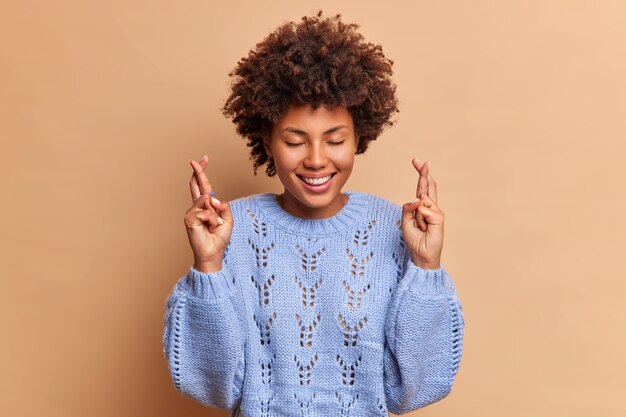 Hopeful beautiful young woman stands with crossed fingers believes dreams come true smiles broadly keeps eyes closed wears knitted sweater isolated over brown studio wall