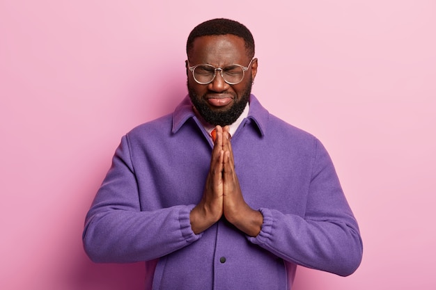 Free photo hopeful black man keeps both hands pressed together in praying gesture, believes in good luck