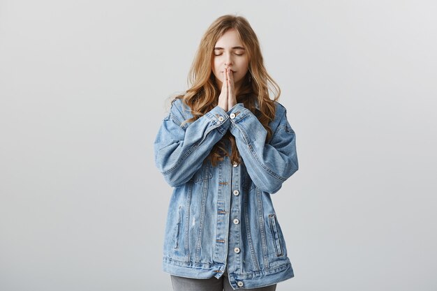 Hopeful blond girl in denim jacket making wish, holding hands in plead, praying