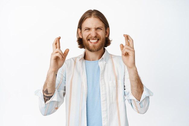 Hopeful blond man, guy having faith, making wish, cross fingers good luck and smiling optimistic, standing in shirt against white background.