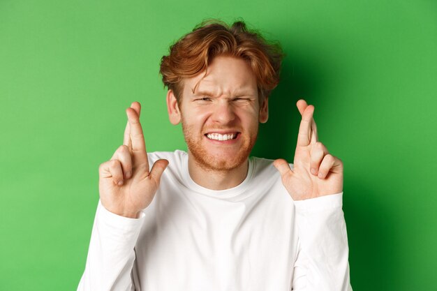 Hopeful caucasian man looking worried, waiting for important results and making wish with fingers crossed, standing over green background