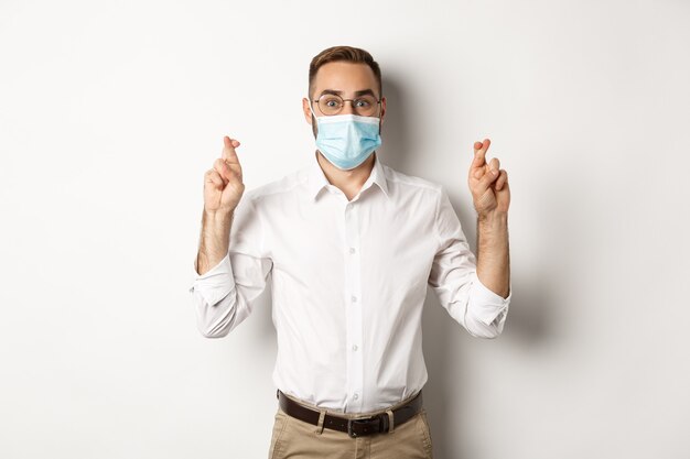   Hopeful employee in medical mask, holding fingers crossed and making wish, white background.