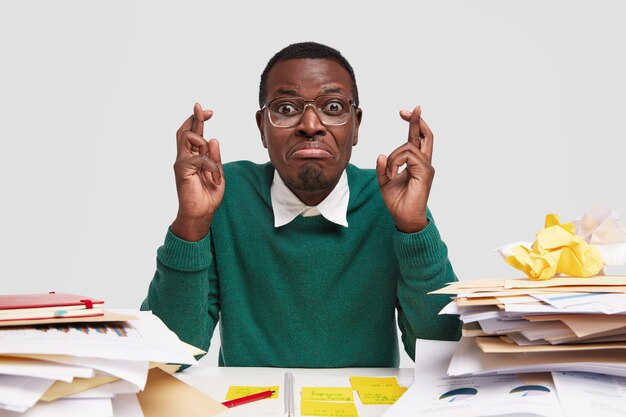 Free photo hopeful miserable student sits at desk with crossed fingers gesture, dressed in casual clothes, wears spectacles, wishes good luck on entrance exam