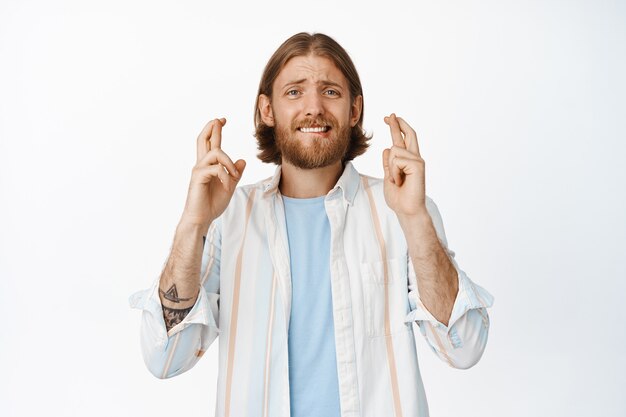 Hopeful nervous bearded guy in shirt, cross fingers good luck, wishing, making wish, anticipating good news, waiting for news, standing on white.