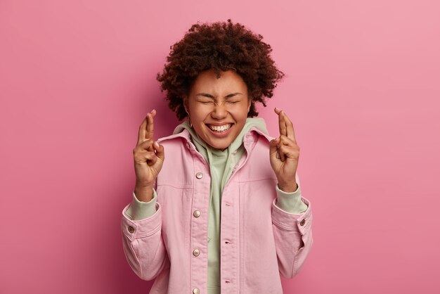 Hopeful optimistic curly woman crosses both fingers, anticipates good news, believes dreams come true, wears hoodie with pink jacket, smiles broadly