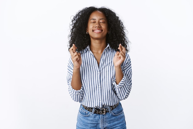 희망에 찬 낙천적이고 매력적인 아프리카계 미국인 여성은 긍정적인 소식을 위해 손가락을 교차합니다.