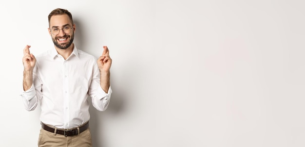 Hopeful optimistic man making a wish cross fingers and praying standing over white background