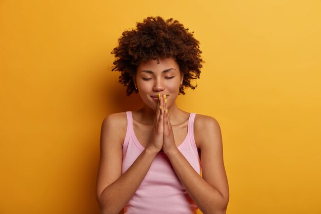 Hopeful pretty female presses palms together in pray, believes in wellness, has big faith, wears casual vest, closes eyes, stays calm, isolated over yellow wall. May my dreams come true.