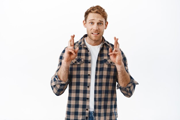 Hopeful redhead adult man cross fingers and looking at camera anticipating making wish praying for dream come true waiting for miracle standing over white background