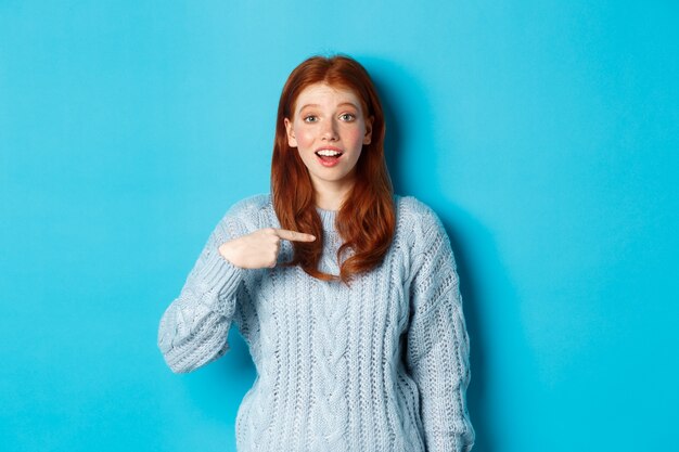 Hopeful redhead girl pointing at herself, standing over blue background