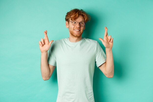 Hopeful redhead man in glasses making a wish, cross fingers and looking right at copy space, dreaming about something or making wish, standing over turquoise background.