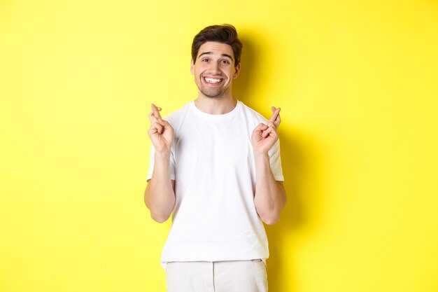 Hopeful smiling man holding fingers crossed, making wish or praying, standing over yellow background.