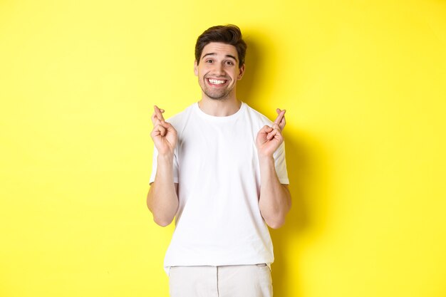 Hopeful smiling man holding fingers crossed, making wish or praying, standing over yellow background