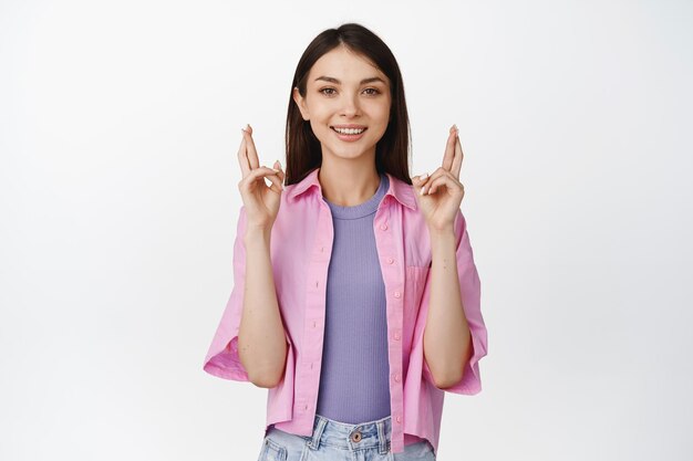 Hopeful teen girl wishes cross fingers for good luck and smiles excited anticipating news standing over white background