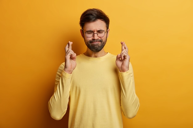 Hopeful unshaven man crosses fingers for good luck, presses lips, has eyes closed, prays for better life, awaits results, wears spectacles and yellow jumper, stands indoor. Monochrome. Hand sign