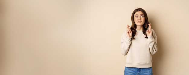 Hopeful worried girl cross fingers making wish standing over beige background