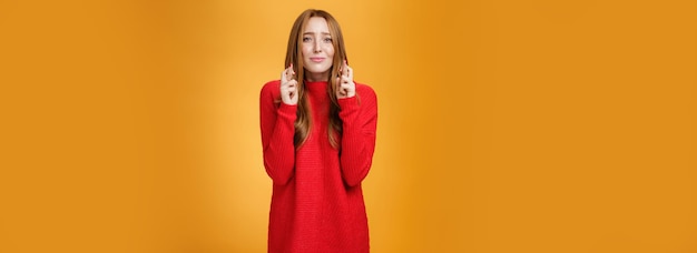 Speranzosa ragazza rossa preoccupata in maglione rosso accigliata che increspa le labbra incrocia le dita per suppli di buona fortuna