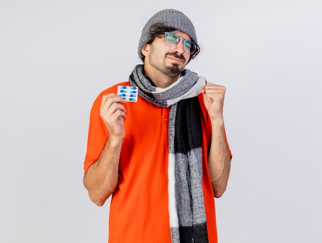Hopeful young caucasian ill man wearing glasses winter hat and scarf holding pack of medical capsules doing be strong gesture with closed eyes isolated on white background with copy space