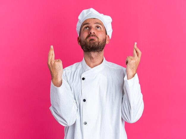 Hopeful young caucasian male cook in chef uniform and cap looking up doing good luck gesture isolated on pink wall