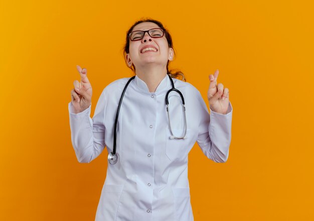 Hopeful young female doctor wearing medical robe and stethoscope and glasses doing crossed fingers gesture wishing for good luck with closed eyes isolated