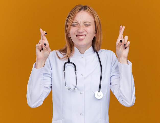 Обнадеживающая молодая женщина-врач-имбирь в медицинском халате и стетоскопе делает жест удачи с закрытыми глазами, изолированными на оранжевой стене