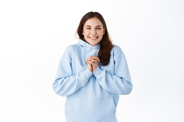 Hopeful young girl student about to receive reward, holding hands clasped near chest, smiling and holding breath, anticipating good news, standing against white wall