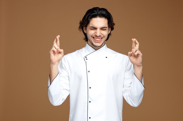 hopeful young male chef wearing uniform making wish crossing fingers with closed eyes isolated on brown background