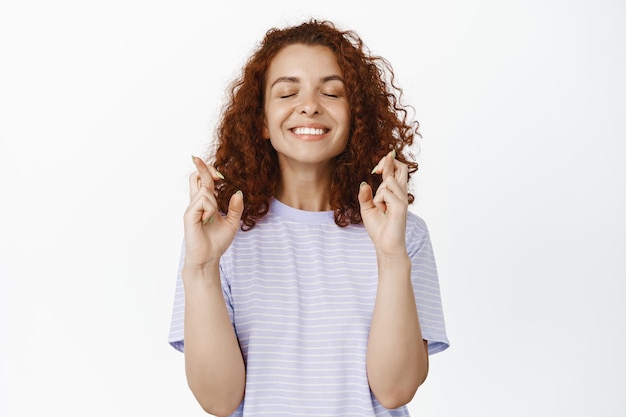 Giovane donna speranzosa con i capelli ricci rossi che esprimono desideri, chiudi gli occhi e incrocia le dita per buona fortuna, augurando, anticipando risultati positivi, sfondo bianco