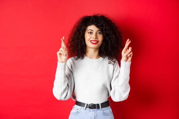 Hopeful young woman with red lips and curly hair, cross fingers for good luck and making wish, praying for dream come true, smiling excited, red background