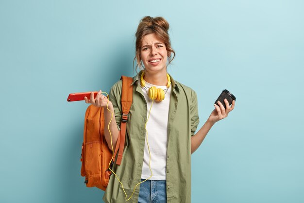 Hopeless dissatisfied woman looks with apathy at camera, upset with softwre problem on mobile phone application, spreads hands in anaware, holds paper cup of drink, has bag on back, smirks face