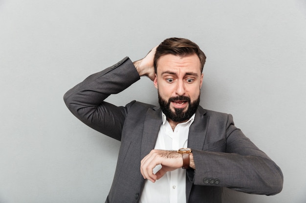 Horizontal astonished man looking at wrist watch, touching his head being late posing isolated over gray