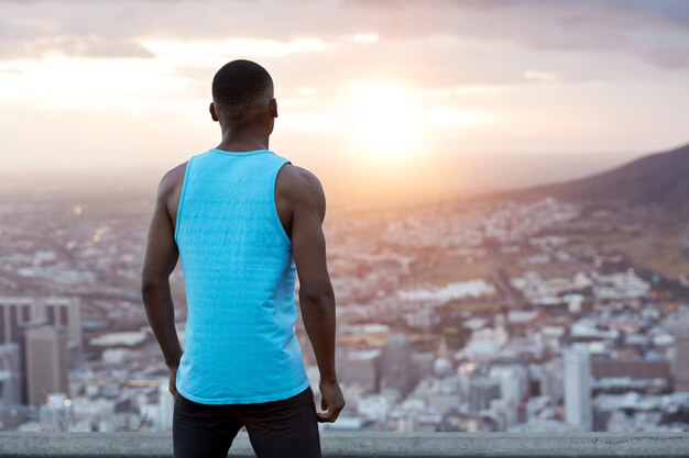 Horizontal back view of athletic man in casual clothes, wears blue vest, takes break after jogging exercise, stands on top in front of magnificent nature view during morning. People, freedom concept