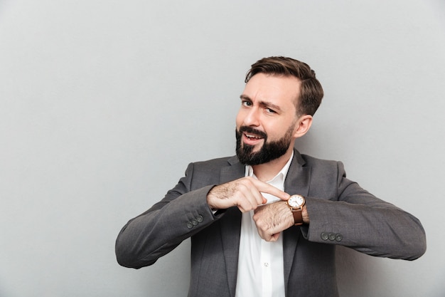 Horizontal bearded man pointing at his wrist watch, posing isolated over gray