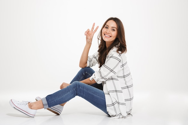 Horizontal cute young woman with brown hair sitting in profile on the floor and showing victory sign, isolated over white wall