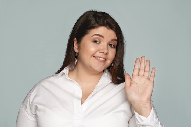 Foto gratuita orizzontale amichevole cercando elegante giovane donna plus size con sorriso allegro agitando la mano, dicendo ciao a te. emozioni positive, segni, gesti e linguaggio del corpo