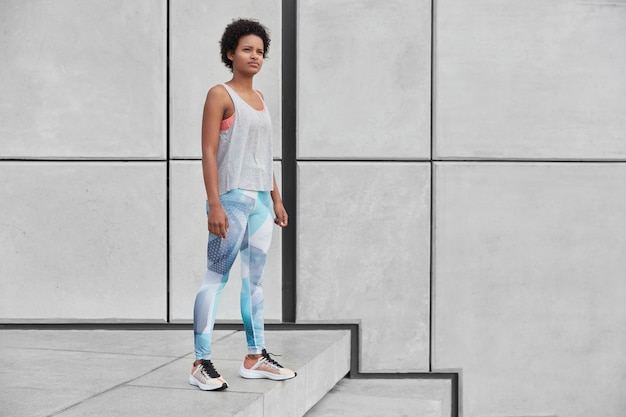 Horizontal full length shot of self determined thoughtful female fitness trainer stands at stairs, wears sportsclothes, going to have gymnastics training with trainee, free space aside for your text