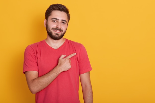 Il maschio con la barba lunga bello orizzontale, vestito in maglietta rossa casuale, indicando da parte il dito indice, mostra lo spazio della copia per la vostra pubblicità o il testo di promozione.